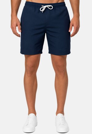 Uomo che indossa pantaloncini da bagno blu navy con coulisse bianca e scarpe da ginnastica bianche, in piedi davanti a uno sfondo chiaro uniforme.