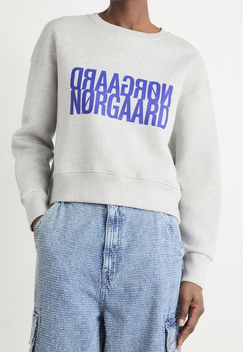 Personne portant un sweat-shirt gris clair avec le texte bleu “NØRGAARD” et un pantalon en denim bleu texturé, visible des épaules aux cuisses.