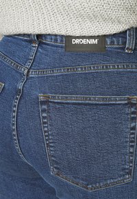Jeans de denim azul escuro com um design clássico de cinco bolsos. Apresenta um rótulo preto da DRDENIM na cintura e costuras visíveis.