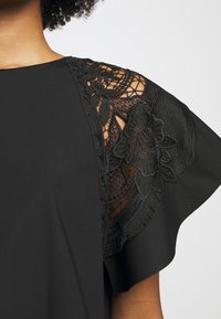Gros plan d'une manche de blouse noire présentant une broderie en dentelle florale et des découpes sur une personne à la peau foncée et aux cheveux bouclés.