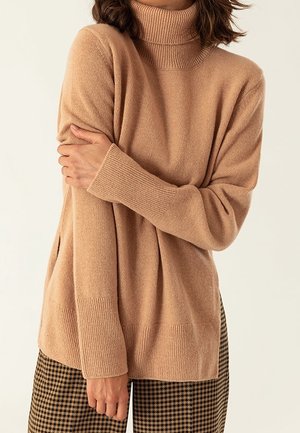 Personne portant un pull à col roulé beige avec des poignets et un bas côtelés, associée à un pantalon à carreaux marron et noir, les bras croisés.