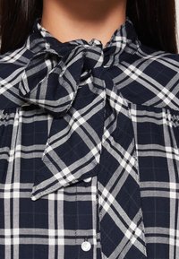 Navy and white checkered blouse with a tie-neck, crafted from woven fabric. Features button closure and a bow accent at the collar.
