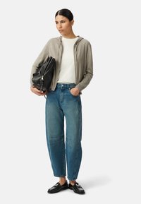 Jasnoszara bluza z zamkiem, biała koszula, niebieskie jeansy z wysokim stanem i szerokimi nogawkami, czarna skórzana torba i czarne loafersy, stojące na neutralnym tle.