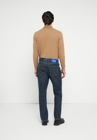 Barna hosszú ujjú ing, sötétkék farmernadrággal párosítva. A farmernadrág derékpántján kék HUGO címke található, egyenes szárú fazonban.