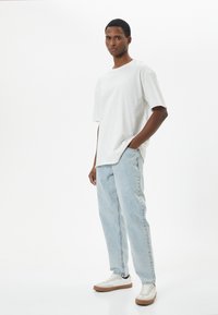 Camiseta blanca de manga corta con un corte relajado, combinada con jeans de mezclilla azul claro. El modelo lleva zapatillas blancas con suelas marrones.