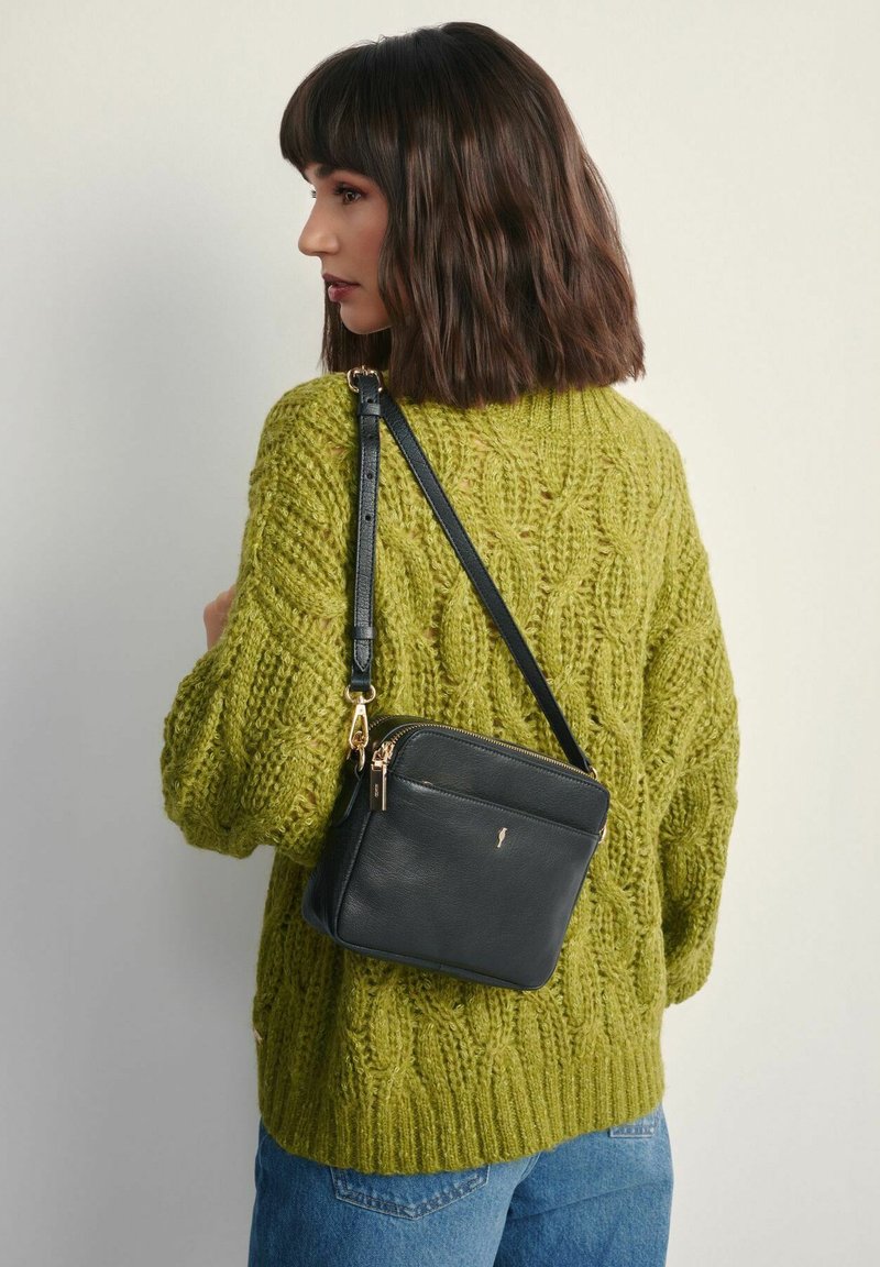 Czarna skórzana torebka typu crossbody z złotymi akcentami, prostokątny kształt i teksturowana powierzchnia, noszona na oliwkowym swetrze z dzianiny.