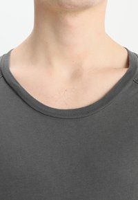 Detailní záběr na krk a horní část hrudníku osoby v tmavě šedém tričku s kulatým výstřihem.