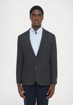 Dunkelgrauer maßgeschneiderter Blazer mit zwei vorderen Taschen, einem einzigen Knopfverschluss und einem blauen Etikettakzent. Dazu wird ein hellblaues, gestreiftes Hemd getragen.