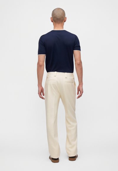 Homme à la tête rasée, de dos, portant une chemise bleu marine à manches courtes, un pantalon crème et des chaussures marron sur fond blanc.