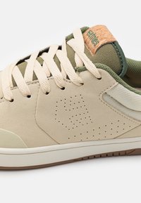 Bežs zamšs sneakers ar olive zaļiem sietiņa akcentiem, krējuma krāsas auklām, korķa piekariņu un perforētu detaļu sānos. Gumijas zole.