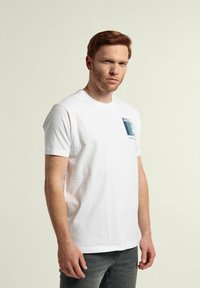 Camiseta de algodón blanca con mangas cortas, cuello redondo, que presenta un estampado gráfico azul en el lado izquierdo del pecho y un diseño minimalista.