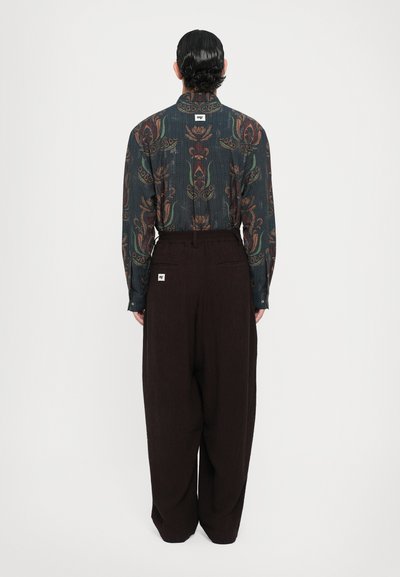 Camicia a maniche lunghe in tessuto scuro con motivi floreali, abbinata a pantaloni larghi marroni. La camicia presenta una texture sottile e un'etichetta all'altezza del colletto.