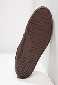 Brun sneaks med texturerad gummisula som har cirkulära mönster. Överdelen har mesh- och släta sektioner, märkta med "LACOSTE."