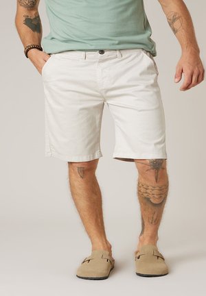 Man draagt een lichtgroen t-shirt, witte knielange shorts, beige instapschoenen, met zichtbare tatoeages op armen en benen.