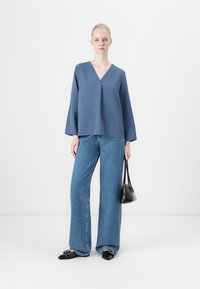 Frau, die eine blaue V-Ausschnitt-Bluse, weite blaue Jeans und schwarze spitze Schuhe trägt, hält eine kleine schwarze Handtasche vor einem schlichten weißen Hintergrund.