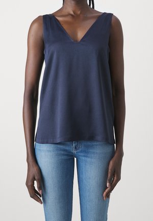 Femme portant un haut sans manches bleu marine à col en V associé à un jean bleu clair, debout devant un fond clair uni.