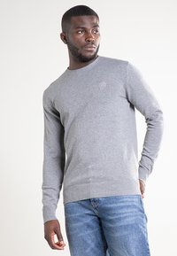 Szary sweter dziany z okrągłym dekoltem, ściągaczami na mankietach i u dołu, z małym wytłoczonym logo na piersi. Noszony z niebieskimi dżinsami.