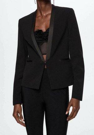 Frau trägt einen schwarzen, maßgeschneiderten Blazer mit Satinrevers über einem schwarzen, floralen Spitzenoberteil und passenden schwarzen Hosen.