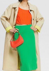 Un trench-coat beige, un crop top orange et une jupe crayon verte sont assortis à un petit sac à main rouge texturé.