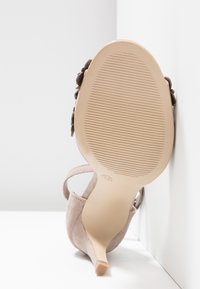 Sandale beige en daim à talon haut fin, avec bride à la cheville et semelle rainurée, inclinée contre un mur blanc d’angle.