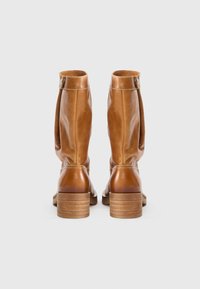 Botas de cuero marrón hasta la rodilla con un diseño holgado, que presentan un tacón apilado y un pequeño detalle de hebilla en el costado. Textura suave y acabado liso.