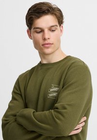Sudadera verde oliva de tela suave, con un cuello redondo y un pequeño diseño de texto gráfico en blanco en el pecho.