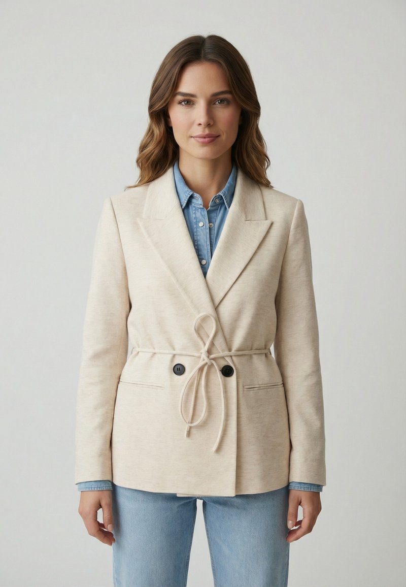 Mujer con cabello castaño claro que lleva un blazer beige con cinturón sobre una camisa de mezclilla azul y vaqueros, posando contra un fondo claro y sencillo.