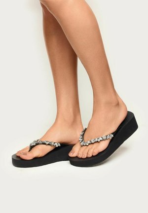 Pies con chanclas negras de plataforma con tiras con joyas sobre un fondo liso y claro.