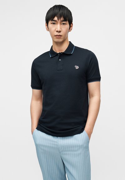 Μπλε ναυτικό πόλο με γιακά, κοντά μανίκια και μικρό κεντημένο λογότυπο στο στήθος. Απαλές γαλάζιες παντελόνες με λεπτές κάθετες ρίγες ολοκληρώνουν την εμφάνιση.