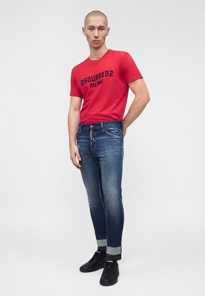 Άνδρας με κόκκινο T-shirt Dsquared2 Milano, τζιν με γυρισμένα μπατζάκια και μαύρα αθλητικά παπούτσια, στέκεται μπροστά σε απλό φόντο.