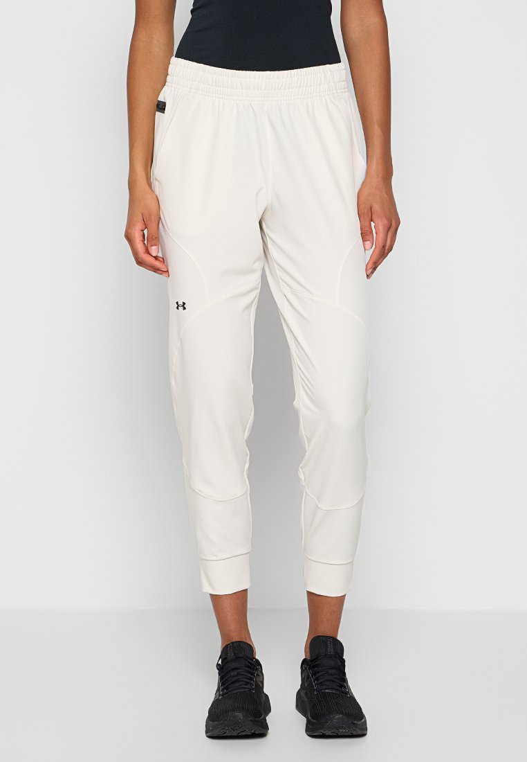Persona vestita con pantaloni jogger bianchi di Under Armour e scarpe da ginnastica nere, in piedi davanti a uno sfondo chiaro e uniforme.