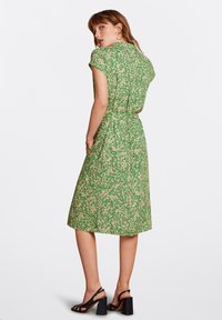 Abito floreale verde con maniche corte, vita elastica e gonna lunghezza ginocchio; presenta un motivo floreale beige chiaro e una texture liscia.