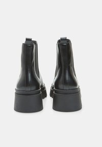 Melnu ādas platformas potīšu zābaki ar elastīgiem sānu paneļiem, skatīšanās no aizmugures uz baltā fona.