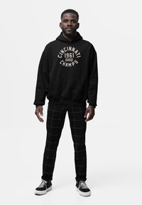 Uomo in piedi, che indossa un hoodie nero con il testo "Cincinnati 1961 Ohio Champs", pantaloni neri con un motivo a griglia bianca e sneakers nere con suole bianche.