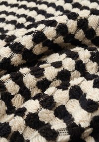 Kohokkuva heittopeitto vaaleassa ja mustassa, jossa on kohotettu pistekuvio. Valmistettu kudotuista kuiduista, joissa on pehmeästä langasta koostuvat vuorotteleva värilohkot.