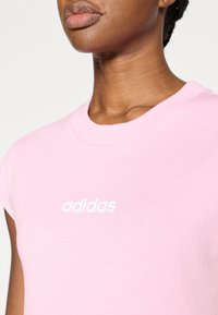 Ružové bavlnené tričko s okrúhlym výstrihom, krátkymi kapkovými rukávmi a bielym logom potlačeným na hrudi. Mäkká textúra a ležérny strih.