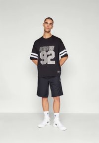 Fekete rövid ujjú póló "CHAMPION 92" grafikával, fehér csíkok a ujjain; fekete rövidnadrággal és fehér tornacipővel párosítva.