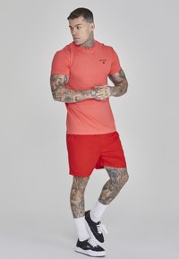 Raudoni sportiniai šortai ir koralų spalvos trumpomis rankovėmis marškiniai. Marškiniai turi logotipą ant krūtinės. Modelis dėvi juodus sportinius batelius ir baltas kojines.
