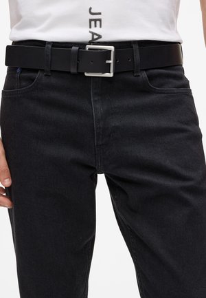 Cinturón de cuero negro con una hebilla rectangular de plata, llevado sobre unos jeans de mezclilla oscuros. La tela es suave con un ligero brillo y mínima costura.