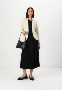 Blazer crème avec col arrondi, associé à une robe noire plissée. Sac à main noir et chaussures en cuir verni avec des embellissements.