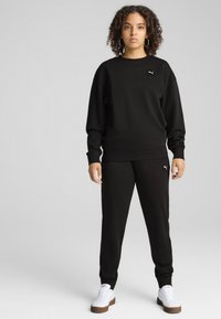Svart sweatshirt och joggers i mjukt tyg. Sweatshirten har en logga på bröstet; joggers har en matchande logga på låret.