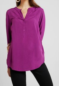 Lila långärmad blus utan krage med knappslå och rundad nederkant, tillverkad av ett slätt tyg.
