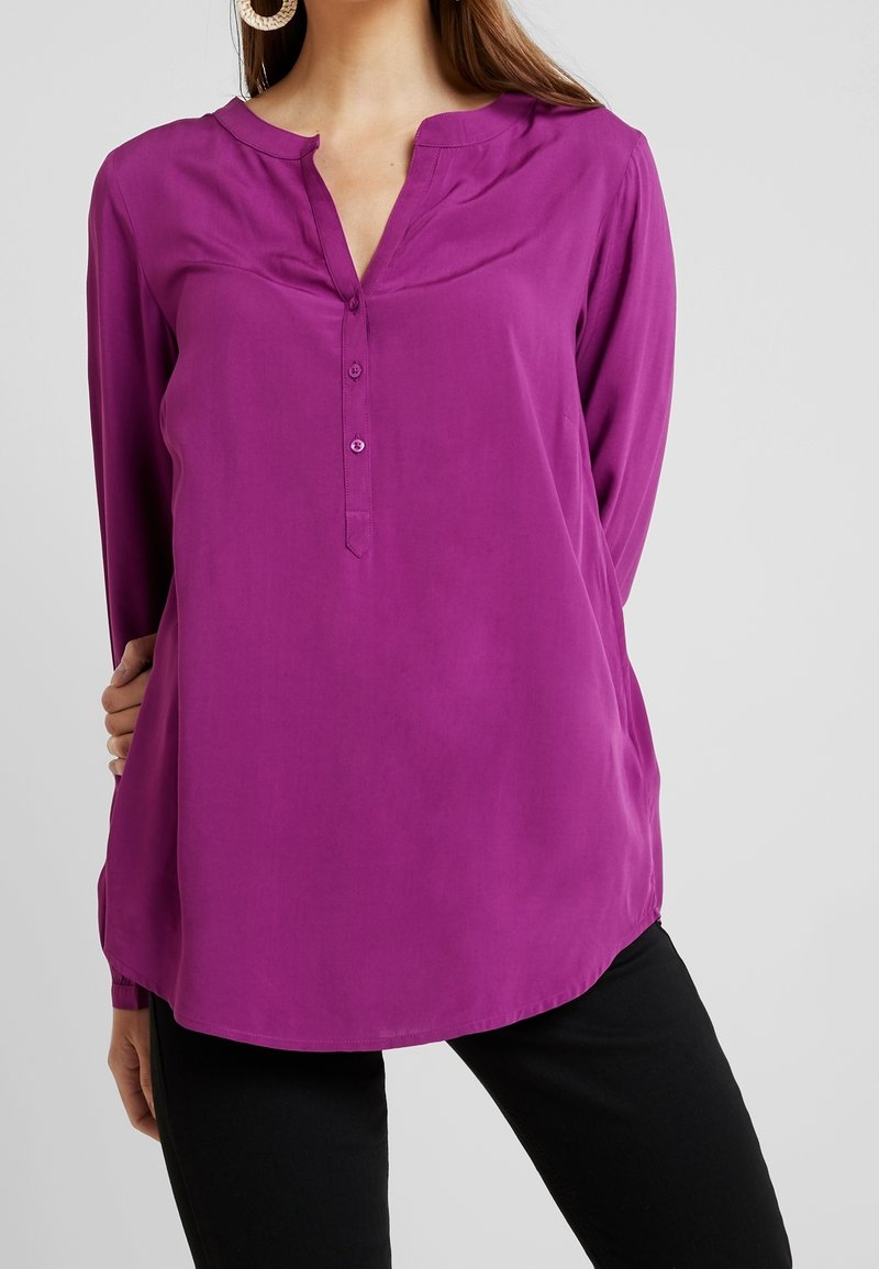Lila långärmad blus utan krage med knappslå och rundad nederkant, tillverkad av ett slätt tyg.