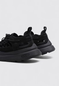 Zapatillas negras con parte superior de malla, collar acolchado y suela de goma gruesa. Las características incluyen un sistema de cordones enredados y una tira de agarre en la parte trasera.