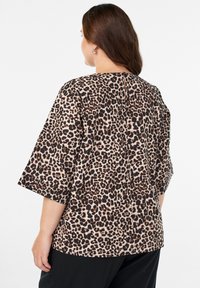 Pikkade pruunide juustega naine, kes kannab hõbedasi rõngaste kõrvarõngaid ja leopardiimuuliga poolte varrukatega tippu, seisab valge taustaga seljaga.