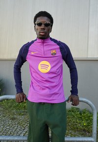 Lilla og marineblå atletisk skjorte med halvlukning, der har et rundt Spotify-logo og et Barcelona-skjold, parret med grønne shorts.