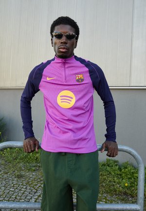 Lilla og marineblå atletisk skjorte med halvlukning, der har et rundt Spotify-logo og et Barcelona-skjold, parret med grønne shorts.