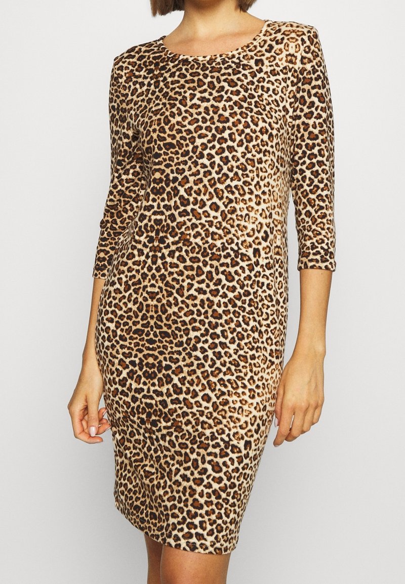 Femme portant une robe ajustée, arrivant aux genoux, avec des manches trois-quarts et un motif léopard noir et marron.