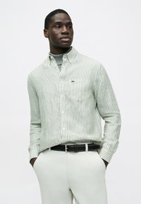 Πράσινο και άσπρο πουκάμισο με ρίγες και κουμπιά, με μικρό λογότυπο, φορεμένο πάνω από γκρι ζιβάγκο, σε συνδυασμό με ανοιχτόχρωμο παντελόνι και μαύρη ζώνη.