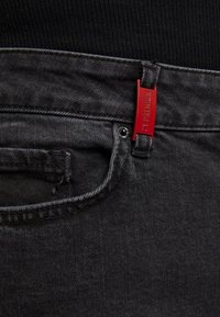 Jeans de mezclilla negros con una etiqueta roja que dice "PREMIUM". Primer plano que muestra los detalles del bolsillo y la costura, con una textura suave y mate.